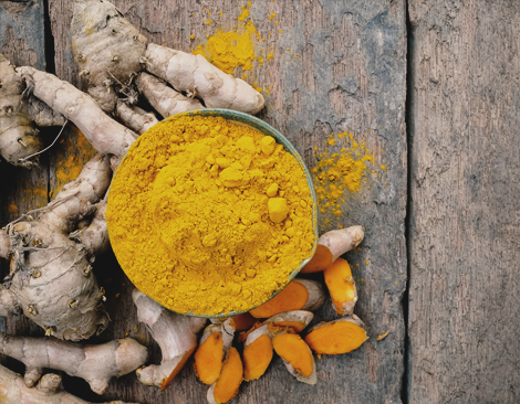 Whole Turmeric roots and a bowl of bright Turmeric powder sit on a wooden surface.
